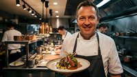 Middle-aged male chef of Caucasian ethnicity wearing white chef's coat and apron, smiling while holding plated dish in professional kitchen with staff working in background, showcasing culinary expertise and restaurant operations.