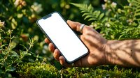 Male hand holding smartphone with blank white screen outdoors in natural green garden setting for mockup and app presentation purposes.