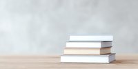 Stack of white and beige hardcover books on wooden table against light gray wall background, minimalist composition for reading and education concept.