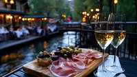 Romantic riverside dining setting with charcuterie board featuring prosciutto, olives and two glasses of white wine on wooden table. Evening ambiance with bokeh lights.