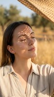 Young woman with long dark hair and fair complexion enjoying sunlight outdoors, wearing beige linen shirt with woven hat creating shadow patterns on face during golden hour.