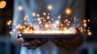 Magical glowing sparkles and light particles floating above hands holding digital tablet in dark room, creating ethereal atmosphere for tech concept.