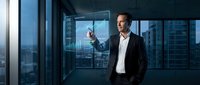 Caucasian male professional in business suit and white shirt standing in modern high-rise office, pointing at digital interface on glass window with cityscape view behind him.