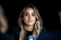 Young Hispanic woman with blonde wavy hair and natural makeup smiling warmly at camera against dark background, wearing navy blue top, professional headshot style portrait.