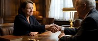 Two mature professionals shaking hands across wooden desk in warm office setting, sealing business agreement or partnership deal with handshake gesture.