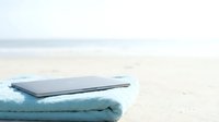 Laptop computer resting on folded towel on sandy beach near ocean water, representing remote work, vacation productivity, digital nomad lifestyle.