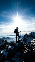 Silhouetted hiker with trekking poles stands on snowy mountain peak against bright sun and blue sky, overlooking vast alpine valley landscape below for adventure and outdoor recreation concepts.