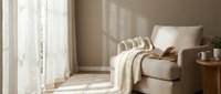 Cozy beige armchair with open book and knit blanket near sunlit sheer curtains in a minimalist neutral living room interior.