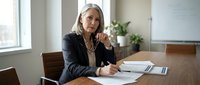 Mature woman with gray hair and light skin tone, wearing professional black blazer, sitting at wooden conference table with notepad and pen, contemplative pose with hand near face, modern office interior with plants and whiteboard visible.