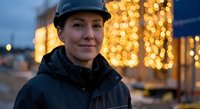 Female construction worker in hard hat smiling at evening job site with warm bokeh lights creating professional industrial atmosphere for safety campaigns.
