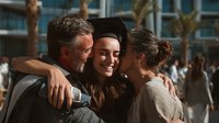 Proud parents embrace their daughter in graduation cap and gown, celebrating academic achievement outdoors with palm trees and modern building in background.