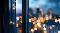 Blurred city lights through window glass creating dreamy bokeh effect at dusk, with dark curtain silhouette adding depth and atmosphere to urban nightscape.
