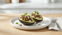 Halved avocado filled with seasoned ground meat and fresh herbs served on white plate in modern kitchen setting for healthy meal preparation and nutritious lunch concepts.