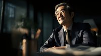 Confident mature Asian businessman in suit smiling while working late at office desk with warm lighting creating professional corporate atmosphere.