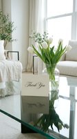 White tulips in a glass vase beside a thank you card on a reflective coffee table in a bright, elegant living room interior.