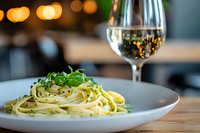 Fresh linguine pasta garnished with arugula on gray ceramic plate, served with white wine glass, restaurant ambiance with soft bokeh lights in background.