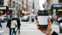 Hand holding blank white coffee cup with black lid against busy urban street with pedestrians and traffic in background, suitable for branding, mockup, and beverage marketing campaigns.