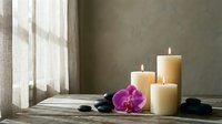 Spa wellness setup with burning candles, purple orchid flower and black stones on wooden surface near window for relaxation and meditation therapy.