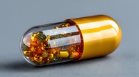 Golden pharmaceutical capsule with colorful medicinal oil beads and particles inside on gray background, suitable for healthcare and supplement advertising.