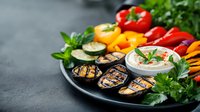 Grilled vegetables and hummus platter with colorful bell peppers, cucumber slices, fresh herbs and roasted eggplant on dark stone background, Mediterranean appetizer.