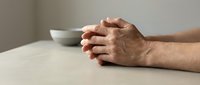 Elderly man hands clasped together in prayer or meditation pose on wooden table with coffee cup in background conveying peace and contemplation.
