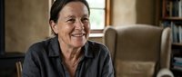 Smiling mature Caucasian woman in casual dark shirt sitting comfortably in cozy home interior with bookshelf background for lifestyle and wellness content.