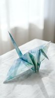 Delicate watercolor origami crane in soft blue and green tones on white fabric surface with natural window lighting for mindfulness and craft projects.