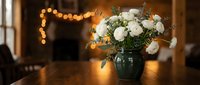 White roses and eucalyptus bouquet in dark vase on wooden table with warm string lights creating cozy romantic atmosphere for home decor and celebrations.