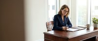 Caucasian woman, middle-aged, wearing dark blazer and glasses, seated at wooden desk reviewing documents in bright office with windows, professional business setting.