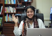 Young South Asian woman wearing headphones smiles and waves during online meeting, sitting at desk with laptop in home office with bookshelf background.