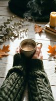 Person with medium skin tone holding steaming mug of coffee on wooden surface surrounded by autumn leaves, eucalyptus branches, candle and knitted sweater for seasonal lifestyle content.