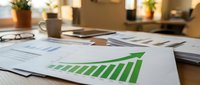 Business growth chart with green upward arrow on desk with financial documents and office supplies in modern workplace setting for corporate success presentations.