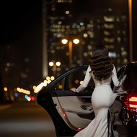 Elegant woman in white dress stepping out of luxury car at night with city lights bokeh background for lifestyle and fashion concepts.