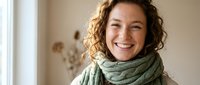 Happy middle-aged woman with curly hair wearing green knit scarf smiling warmly in bright home interior with natural window light for lifestyle content.