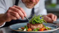 Professional chef garnishing gourmet beef steak with fresh herbs and vegetables on elegant blue plate in restaurant kitchen for fine dining presentation.