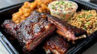 Smoked barbecue ribs with mac and cheese, baked beans and creamy coleslaw served in black container. Tender meat with caramelized BBQ sauce glaze shows perfect smoke ring.