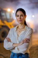 Confident Hispanic woman in white blouse and jeans standing with crossed arms at construction site during sunset, bokeh lights in background, professional portrait.