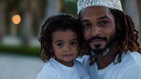 African American father with dreadlocks wearing crown poses with young child outdoors at dusk, both in white shirts, creating heartwarming family portrait.