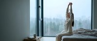 Woman stretching in bed by large window overlooking misty forest landscape, morning wellness and relaxation routine for lifestyle and home decor use.