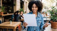 Young African American woman reading document with smile at outdoor cafe terrace. Happy female entrepreneur reviewing business papers at restaurant patio.