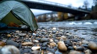 Green camping tent pitched on rocky riverbank with flowing water and bridge in background, moody outdoor adventure scene captured in soft natural lighting.