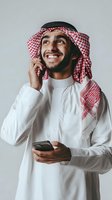 Young Middle Eastern businessman in traditional white thobe and red checkered keffiyeh talking on phone while holding smartphone, smiling and looking up.