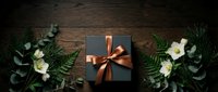 Elegant black gift box with copper ribbon surrounded by ferns and white flowers on dark wood, perfect for holiday and luxury gifting concepts.