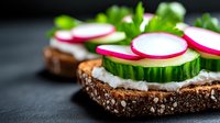 Fresh sandwich with cream cheese, sliced cucumber, radish and herbs on dark rye bread against black background. Healthy vegetarian appetizer.