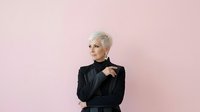 Mature woman with short platinum blonde hair wearing black blazer against pink background, studio portrait for business and professional use.