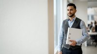 Young Middle Eastern male professional in business casual attire holding laptop, standing in modern office corridor with natural light, suitable for corporate, technology, and business concepts.