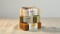 Stack of three natural handmade soaps with botanical labels and dried lavender on top, displayed on wooden surface for organic skincare and wellness product marketing.