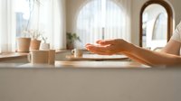 Woman's hands gesturing over a bright minimalist kitchen counter with mugs and soft natural light, creating a calm domestic atmosphere.
