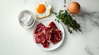 Raw beef steaks on white plate with fresh thyme sprigs, fried egg and whole egg on luxurious marble countertop, overhead view of premium meat preparation.