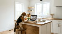 Young woman with long brown hair working from home on laptop computer in modern white kitchen with wooden countertop during video conference call.